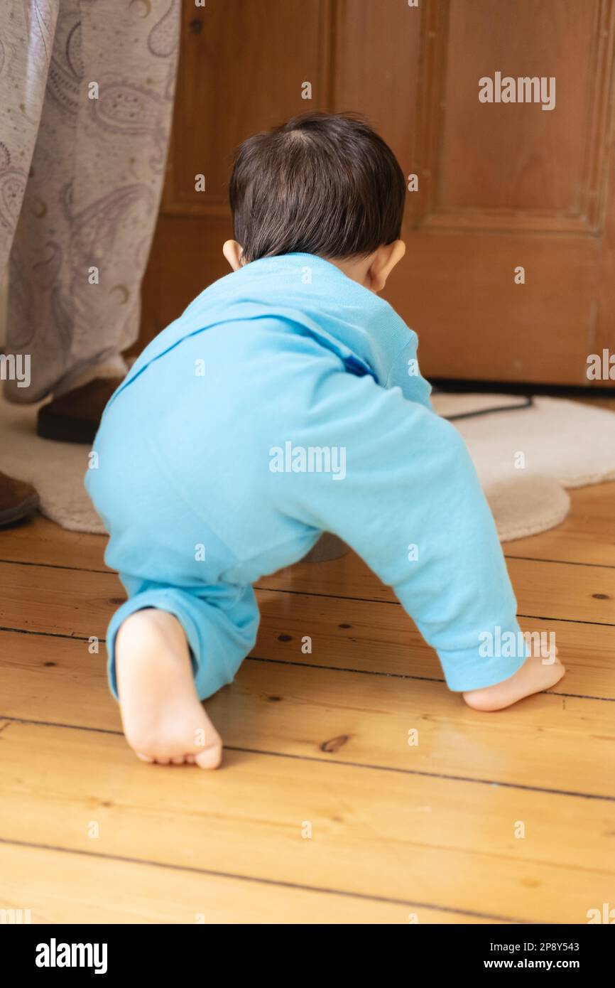 An unrecognizable male multiracial Malaysian and Spanish baby wearing blue pajamas crawling and walking way on wooden floor at home. Feet and barefoot Stock Photo