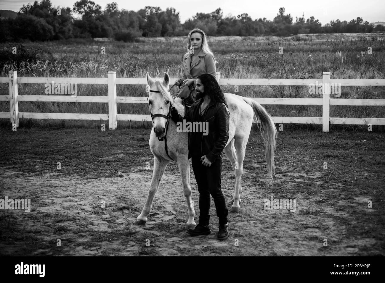 Couple walking with horse on countryside. Handsome bearded man driving horse for a bridle and looking at his girlfriend. Woman riding grey arabian Stock Photo