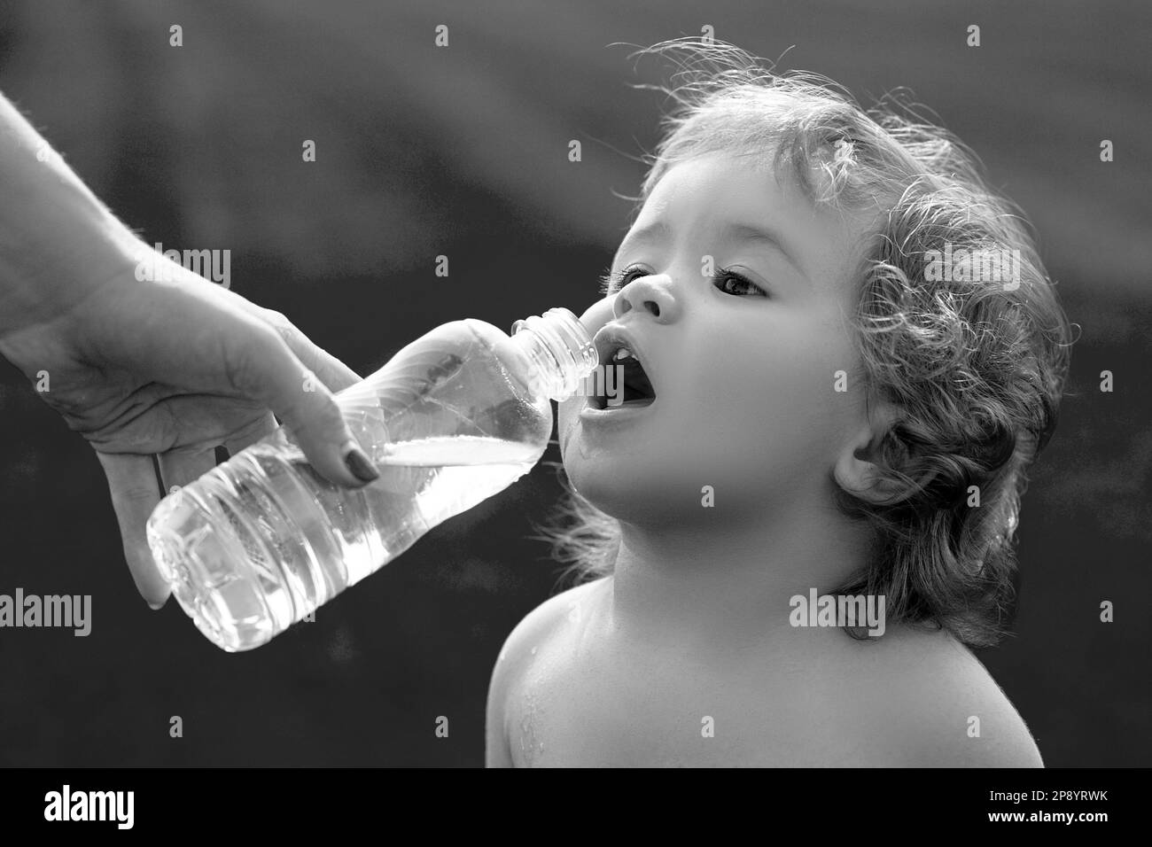 Baby drinking water from mother hands. Child drinks water from a bottle while walking on grass field, baby health. The mother gives the child a bottle Stock Photo