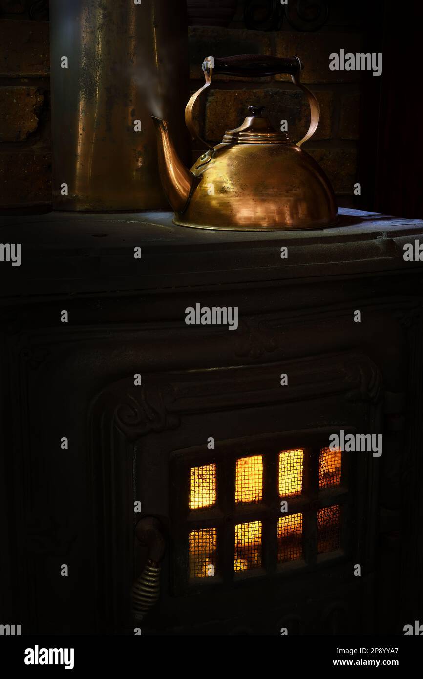 A shiny Copper kettle with steam coming out, sat on an old Cast-iron Wood-burning Stove with a yellow and orange flame glow, in soft, dark mood lighting Stock Photo