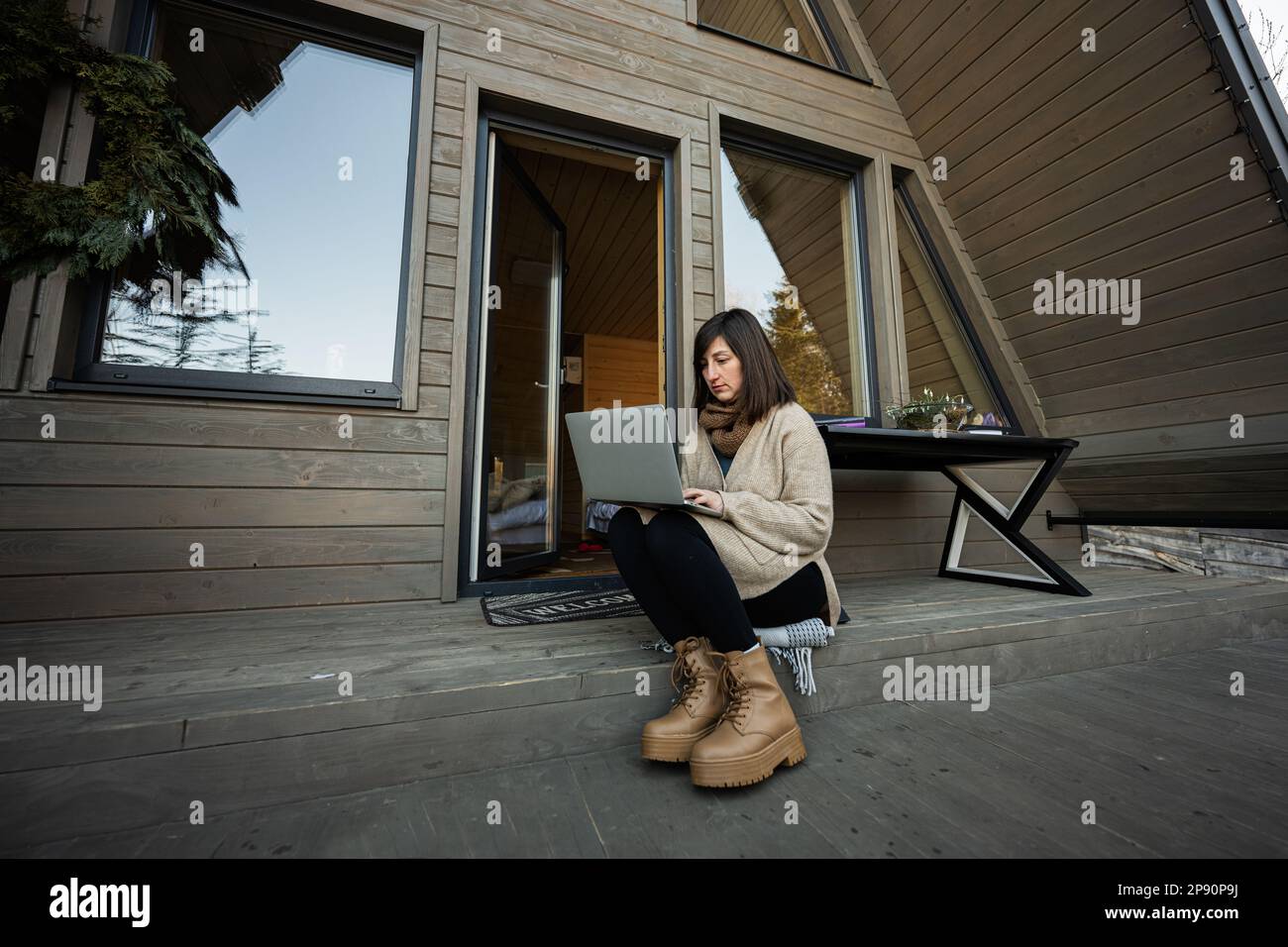 Remote work and escaping to nature concept. Woman works on laptop against tiny cabin house. Stock Photo