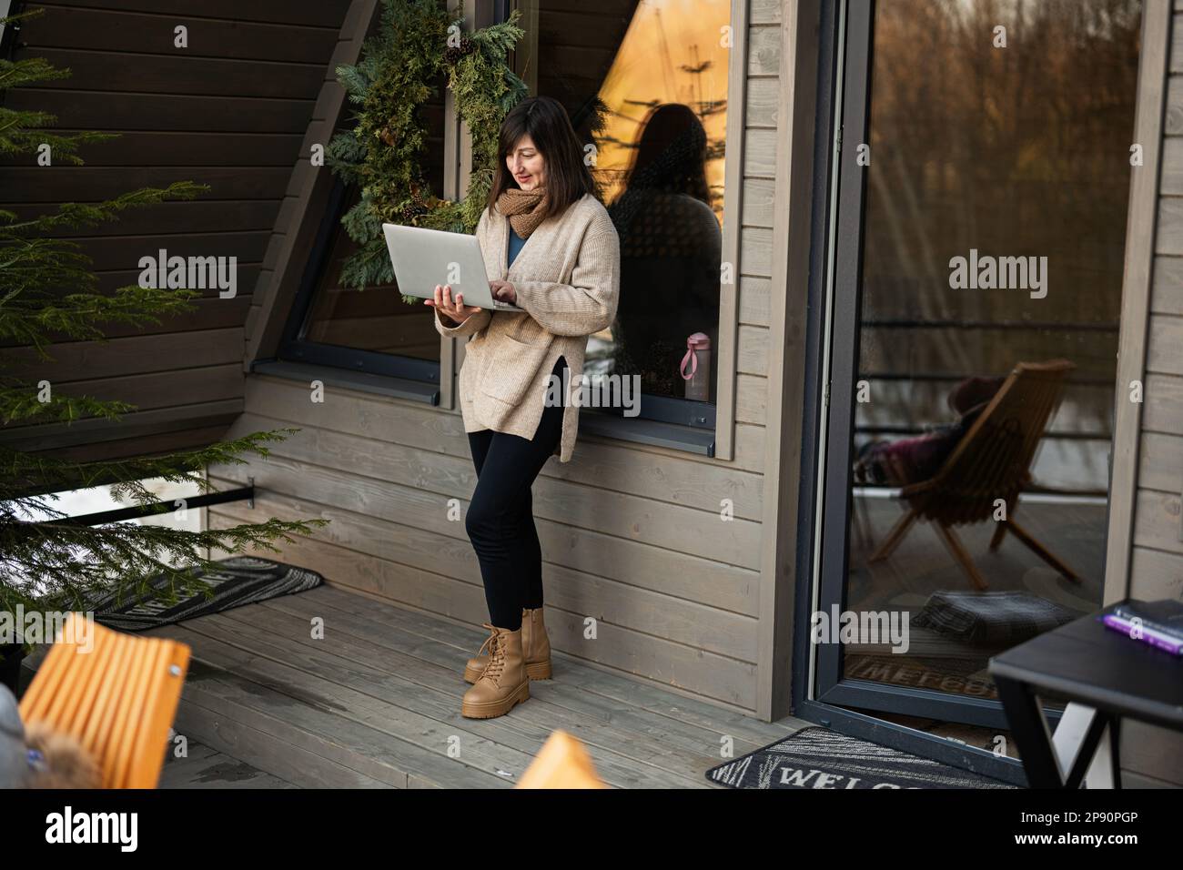 Remote work and escaping to nature concept. Woman works on laptop against tiny cabin house. Stock Photo