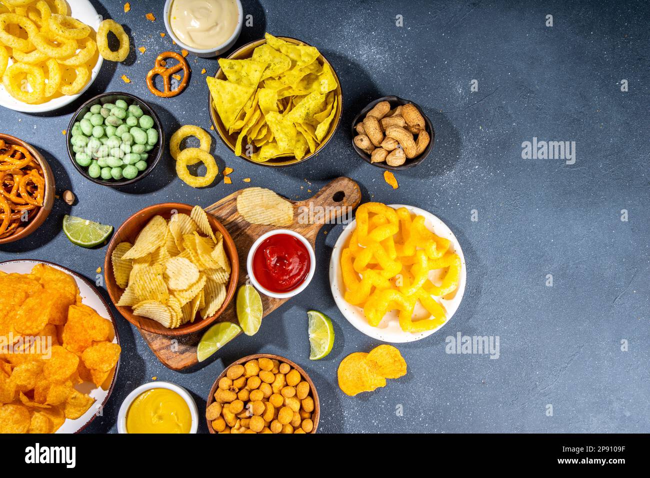 Various unhealthy salty bar snacks set. Dark grey table background with traditional party snacks - chips, onion rings, salted nuts, crisps,  pretzels Stock Photo