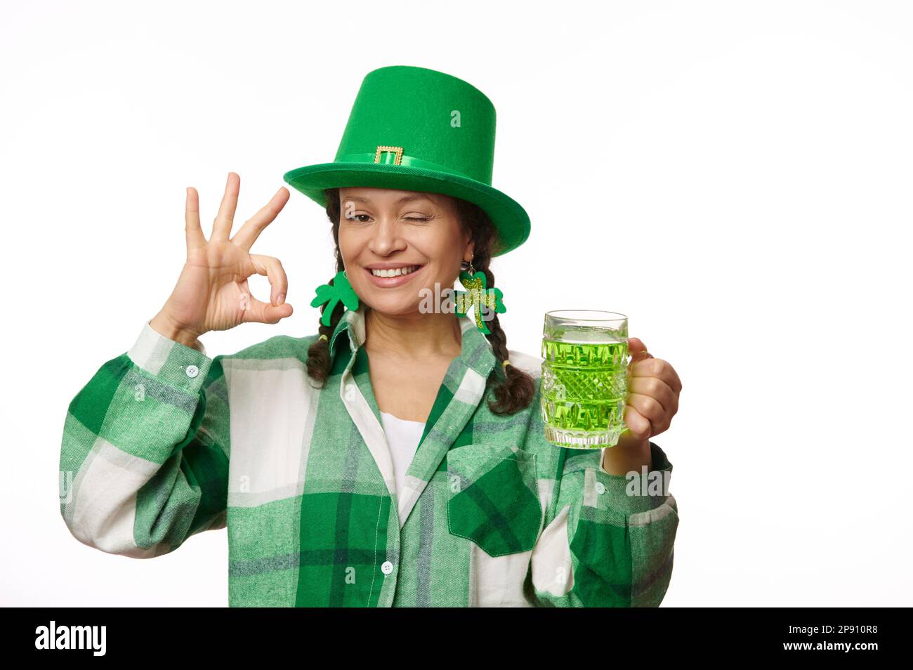 Cheerful woman in green party hat, holds a mug of Irish beer, winks, shows Ok hand sign at camera. Saint Patrick's Day Stock Photo