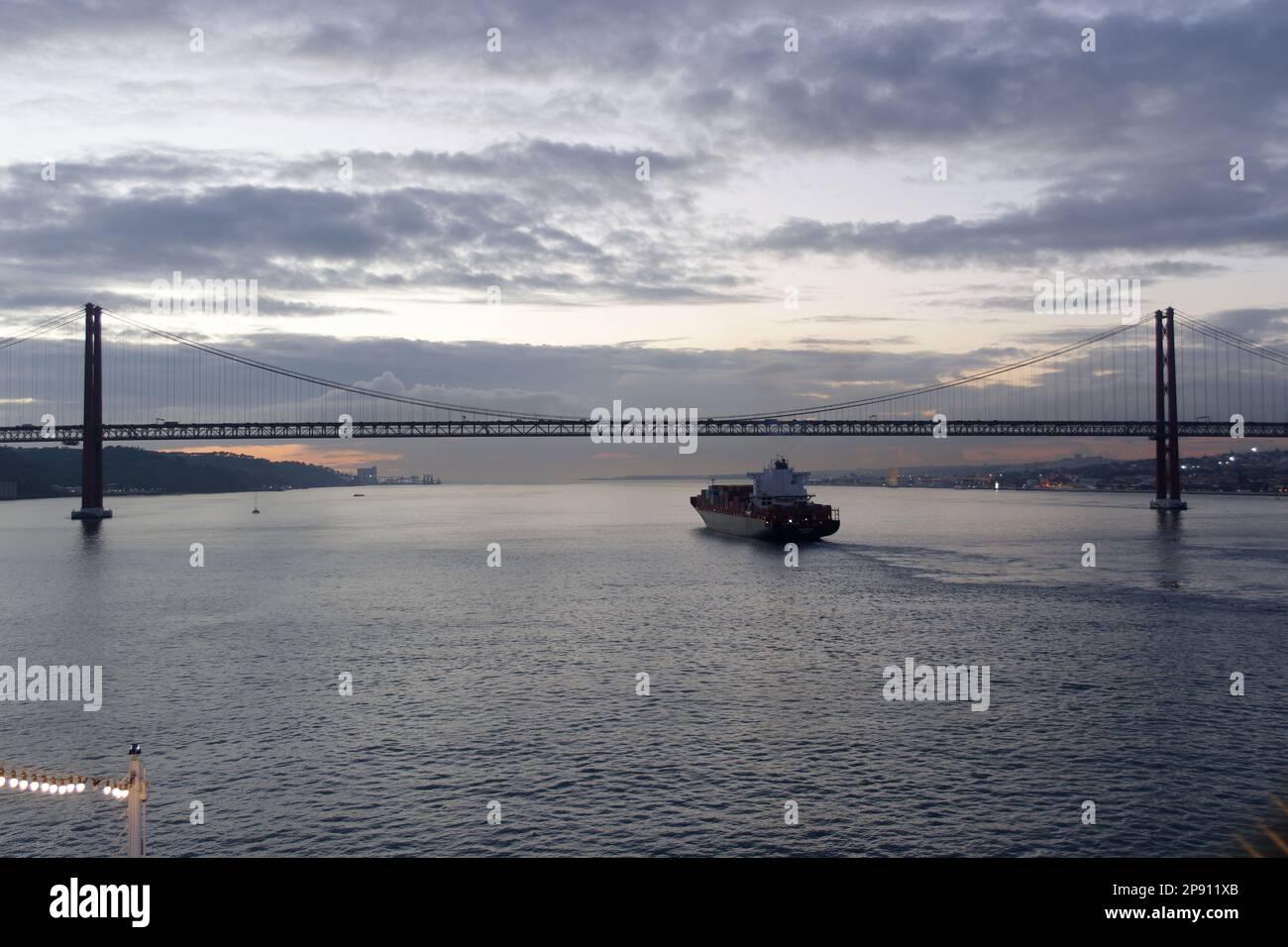 Cargo Ship heading out to sea under the 25 de Abril Bridge in Lisbon, Portugal. In The early evening. Stock Photo