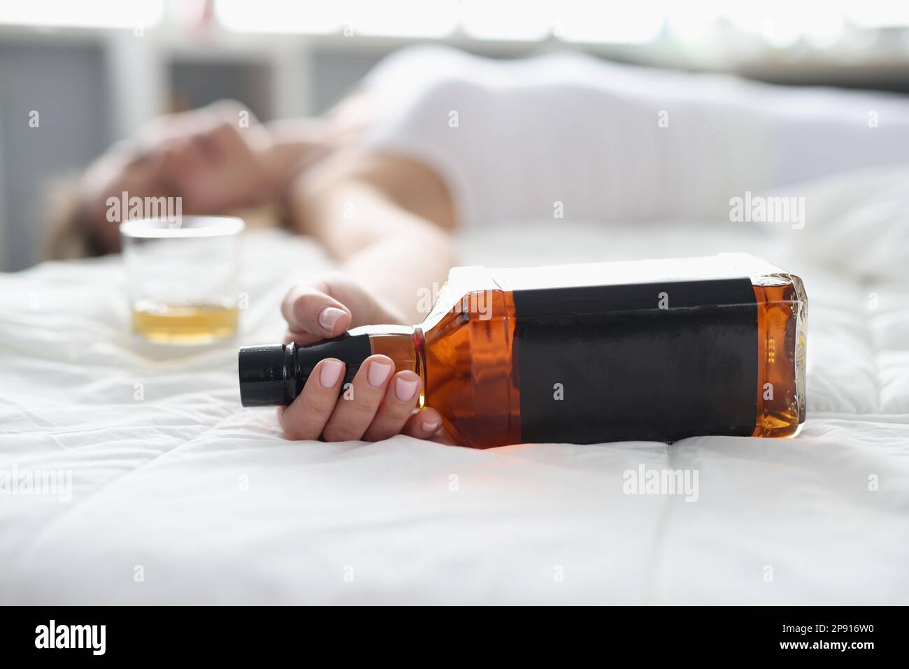 Woman lies in white bed with bottle of whiskey in hand Stock Photo