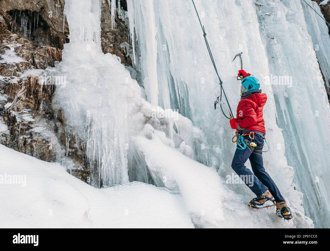 Ice climber dressed in warm winter climbing clothes, safety harness and helmet climbing frozen waterfall using two Ice climbing axes and crampons. Act Stock Photo