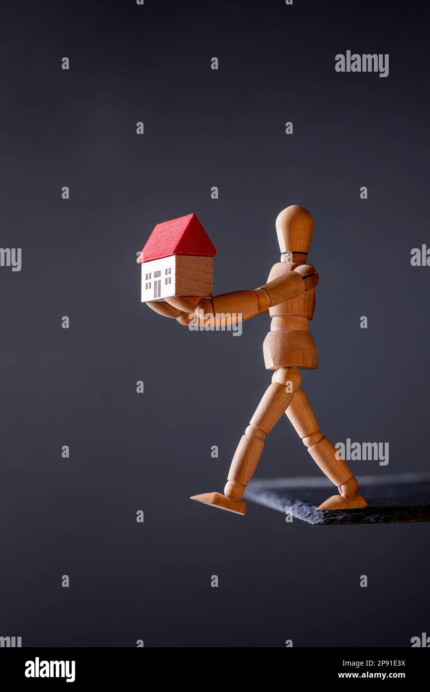 A wooden manikin carries a house while threatening to fall off the edge of a stone slab Stock Photo