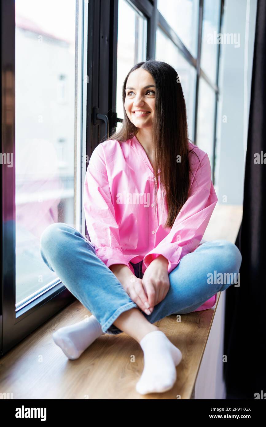 Pretty young woman sitting on windowsill relaxing at home looking at camera. Smiling calm lady chilling in apartment, dreaming, thinking of peaceful t Stock Photo