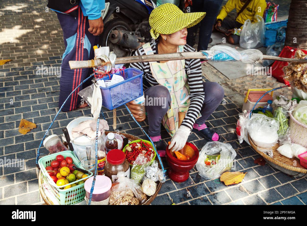 Pattaya, Thailand - March 24, 2016: Asian woman sitting on a sidewalk, cooking and selling traditional Thai meal. Street food vendor with baskets Stock Photo