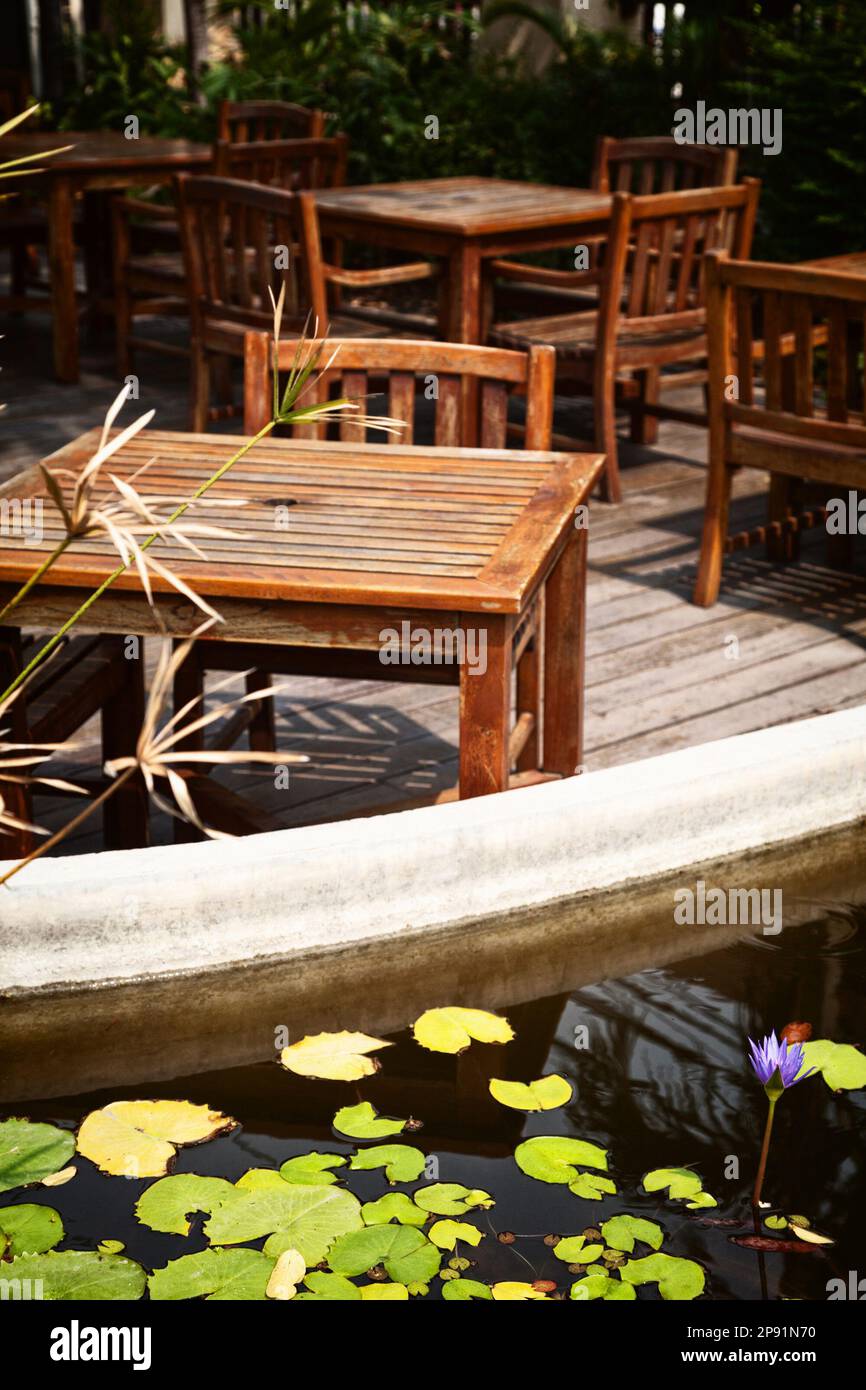 Outdoor cafe patio with old, shabby wooden tables and chairs in the sunlight. Pond with water lilies on a  romantic restaurant terrace Stock Photo