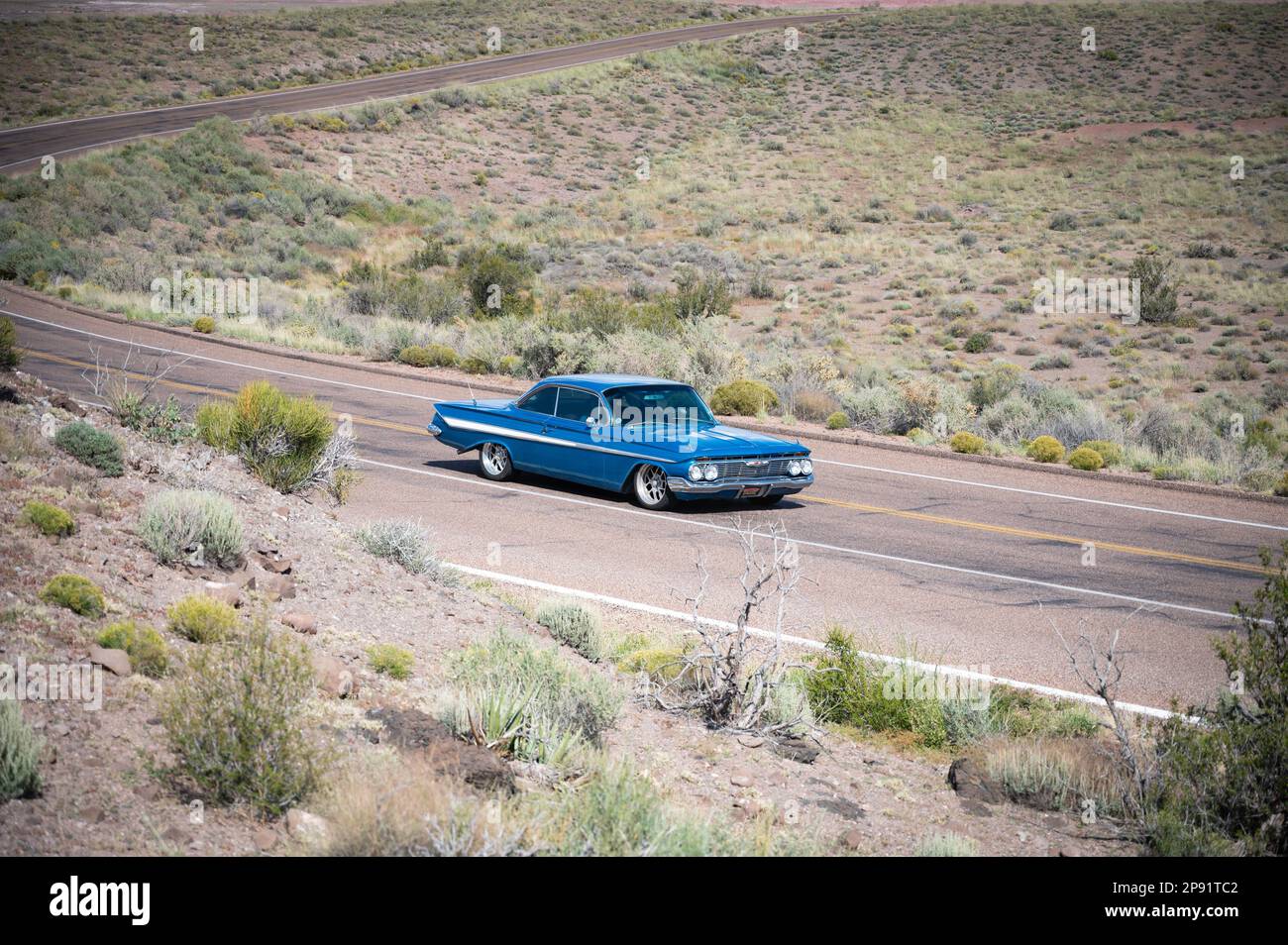 A classic blue car is driving down a desert highway, with hills visible ...