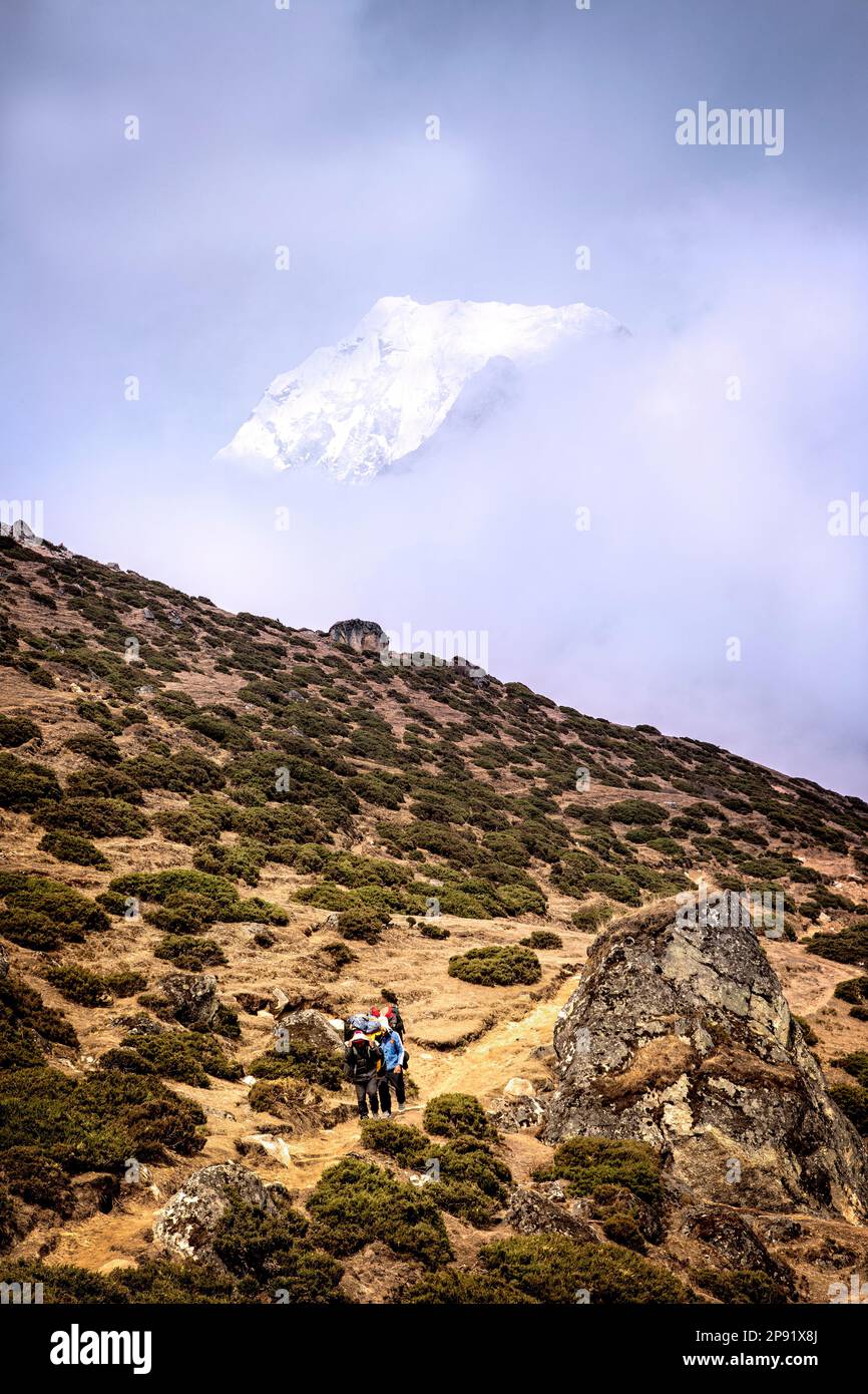 Hikers on the trail to Macchermo with Cholatse appearing through the clouds.  Sagarmatha National Park, Nepal. Stock Photo