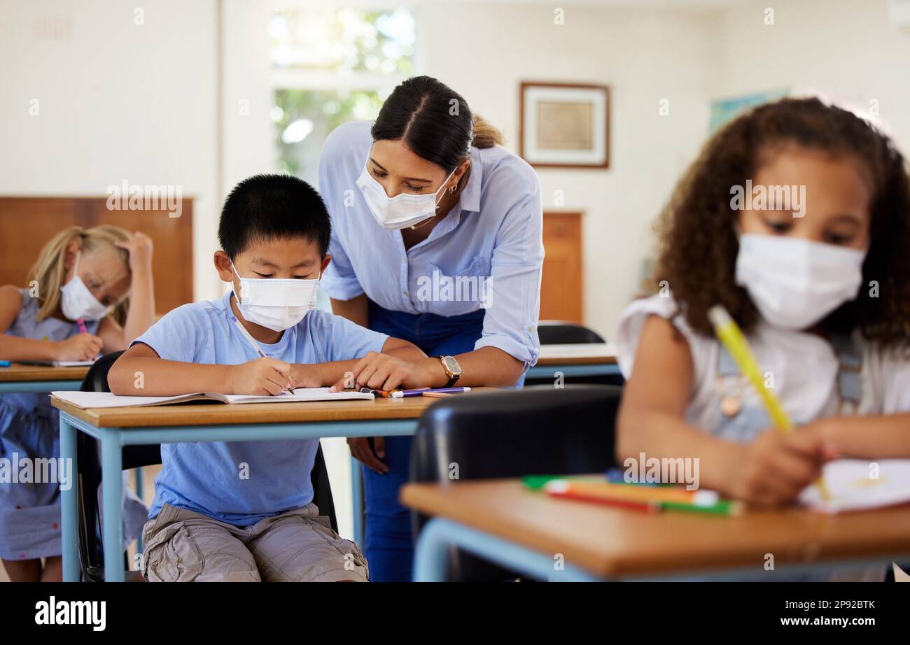 Covid, education and learning with a teacher wearing a mask and helping a male student in class during school. Young boy studying in a classroom with Stock Photo