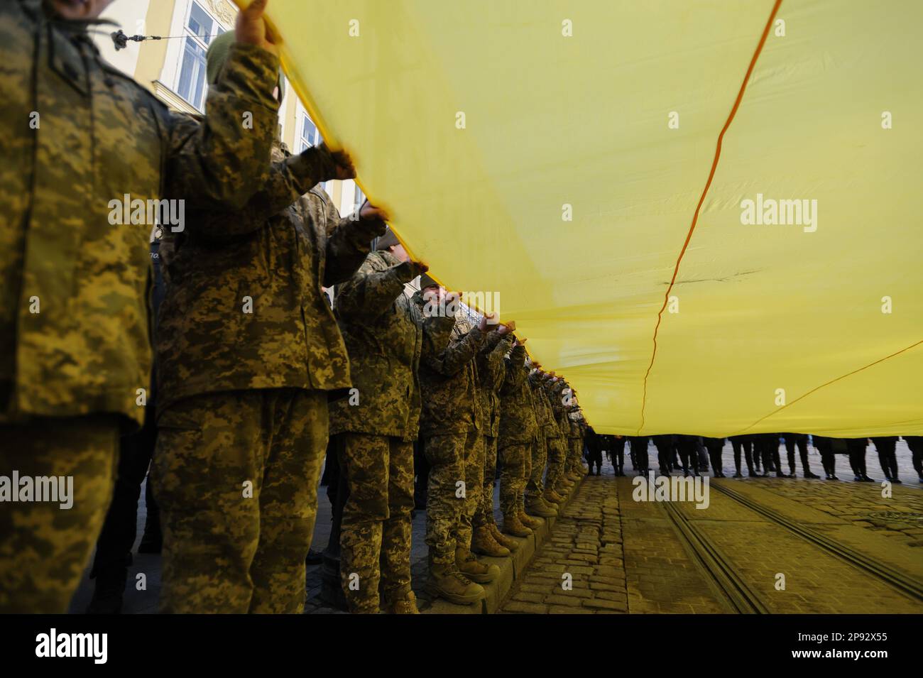 Lviv, Ukraine. 10th Mar, 2023. Military personnel, cadets and policemen sing the national anthem while holding a huge Ukrainian flag in front of the city hall Ratusha in honor of the anniversary of the first performance of the national anthem 158 years ago. Credit: SOPA Images Limited/Alamy Live News Stock Photo