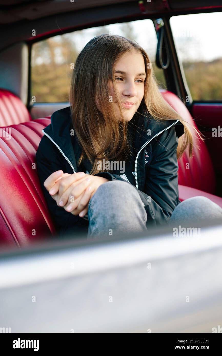 Girl sitting smiling in old car with red leather seats and looking into the distance Stock Photo