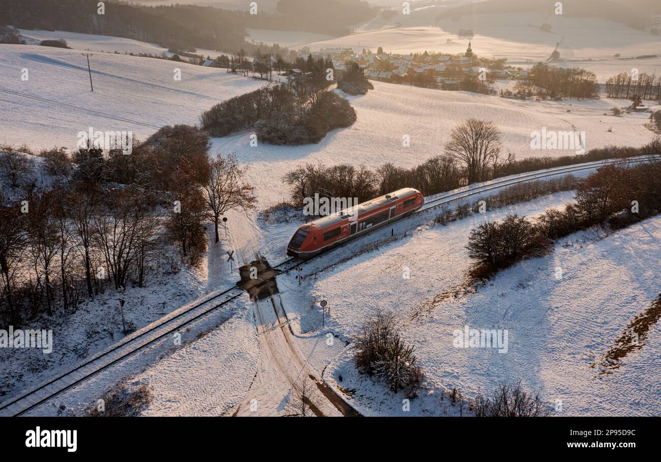 Germany, Thuringia, Allendorf (background), regional train 60, train 29887, level crossing, country lane, landscape, village, fields, forest, snow, overview, oblique view, evening light, back light Stock Photo