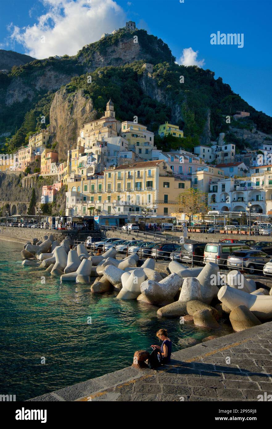 Person sitting on the main jetty at Amalfi, Salerno, Italy  with the town rising, clinging to the hillside. Amalfi Coast. Tyrrhenian Sea. Stock Photo
