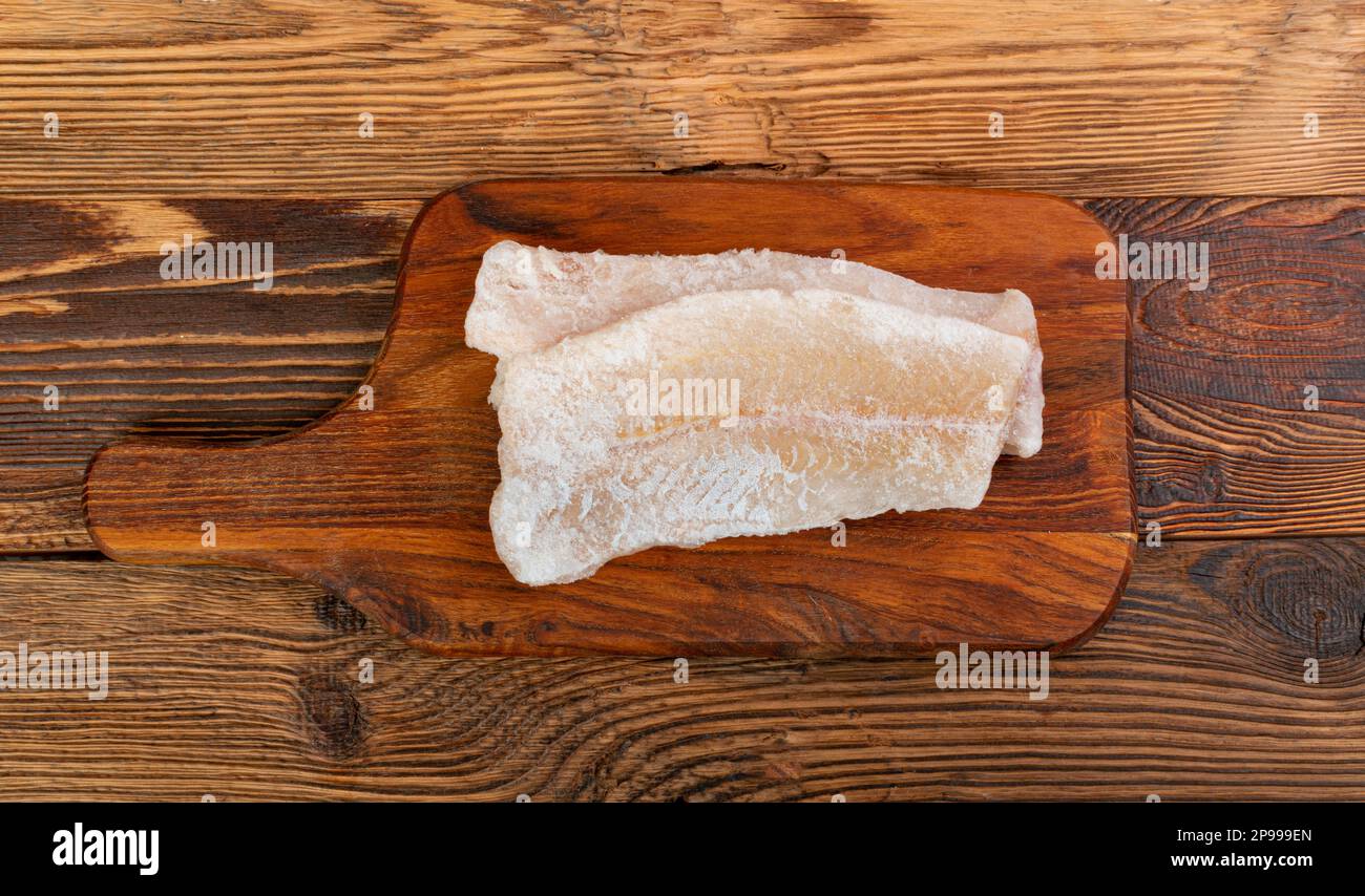 Frozen Fish on Wooden Cutting Board, White Cod Fillet, Iced Hake Filet, Frozen Pollock Meat on Rustic Wood Background Top View Stock Photo