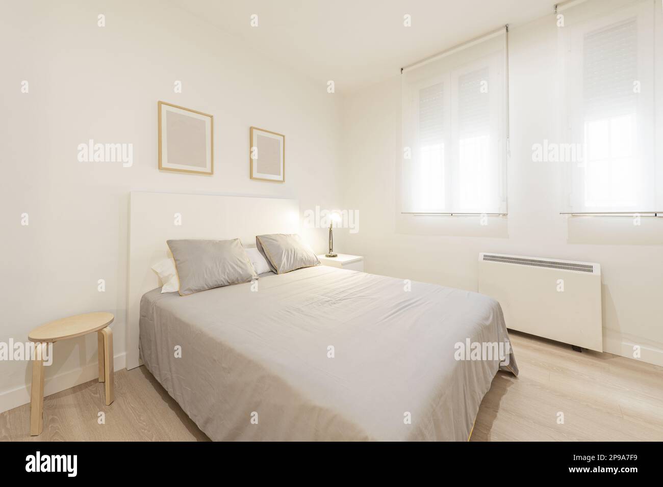 Bedroom with a double bed with wooden bedside tables, a gray bedspread with cushions, double windows with roller shutters and an electric heater Stock Photo