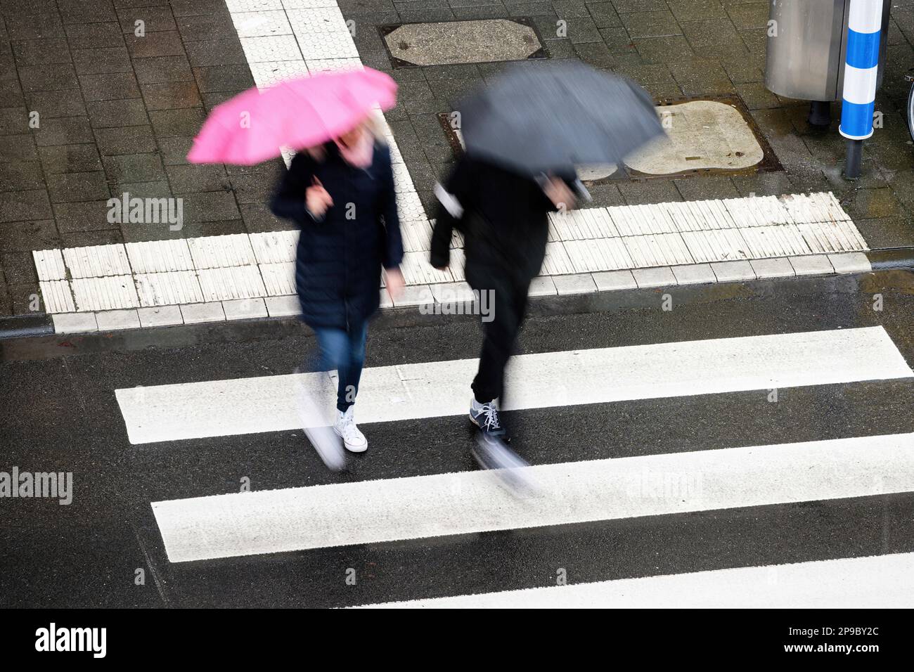 motion blur of two pedestrians with black and pink umbrellas crossing a ...