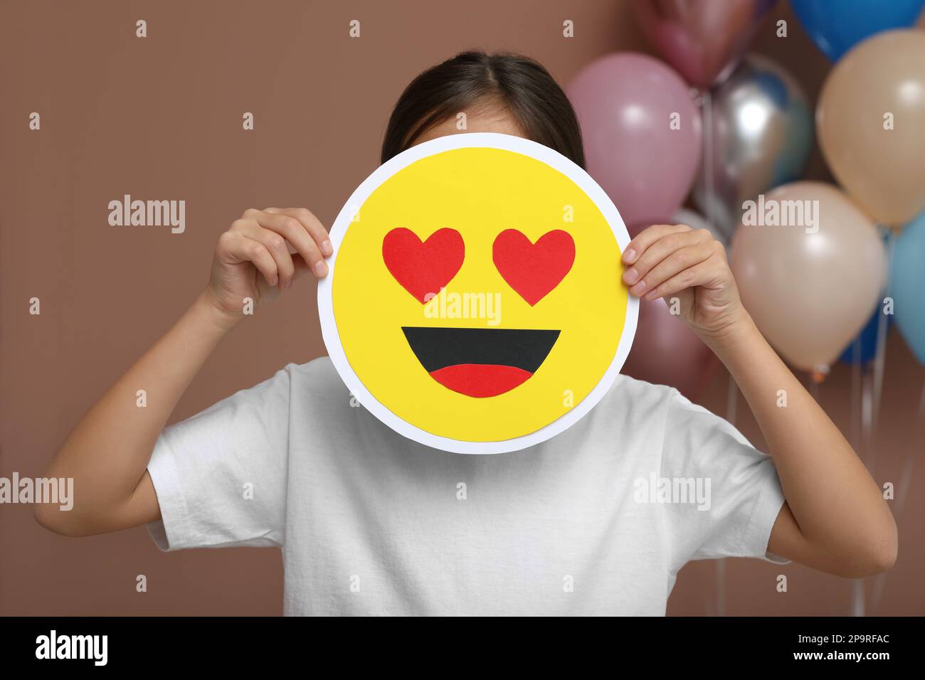 Little girl covering face with heart eyes emoji in decorated room Stock