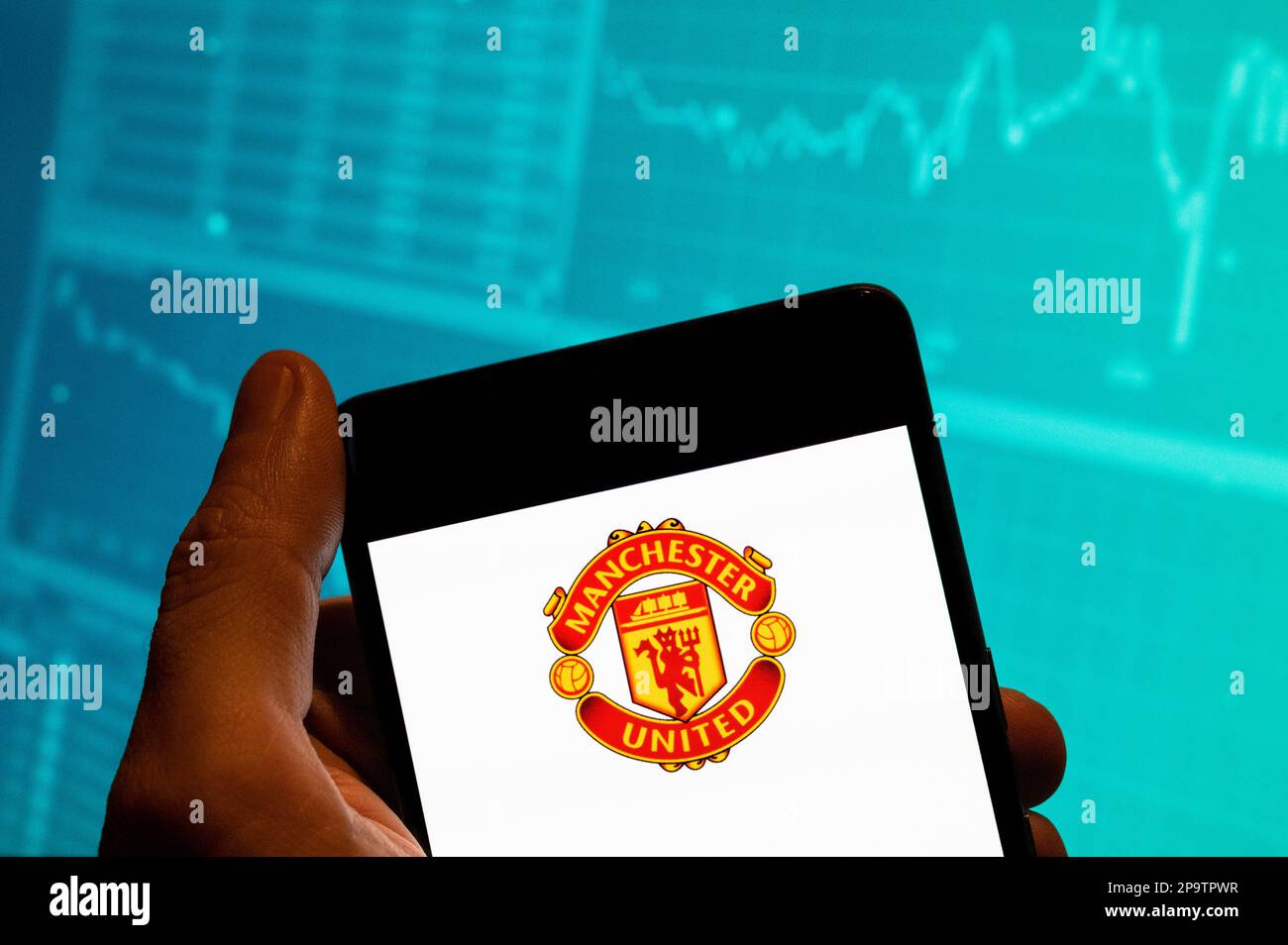 In this photo illustration, the professional premier league football club Manchester United (MU) logo is seen displayed on a smartphone with an economic stock exchange index graph in the background. Stock Photo