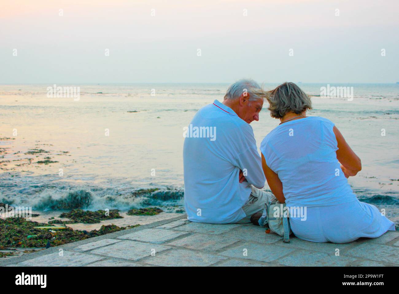 Old elderly couple seated by the sea shore / on the beach at sunset ...