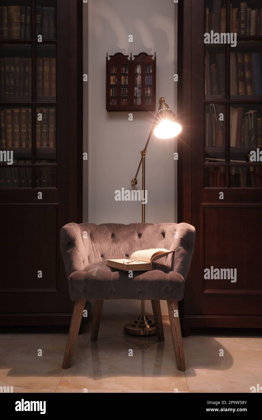 Comfortable armchair with book and lamp between wooden bookcases in library Stock Photo