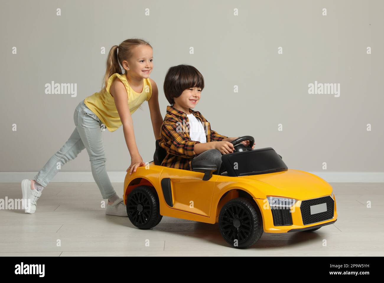 Cute girl pushing children's electric toy car with little boy near grey