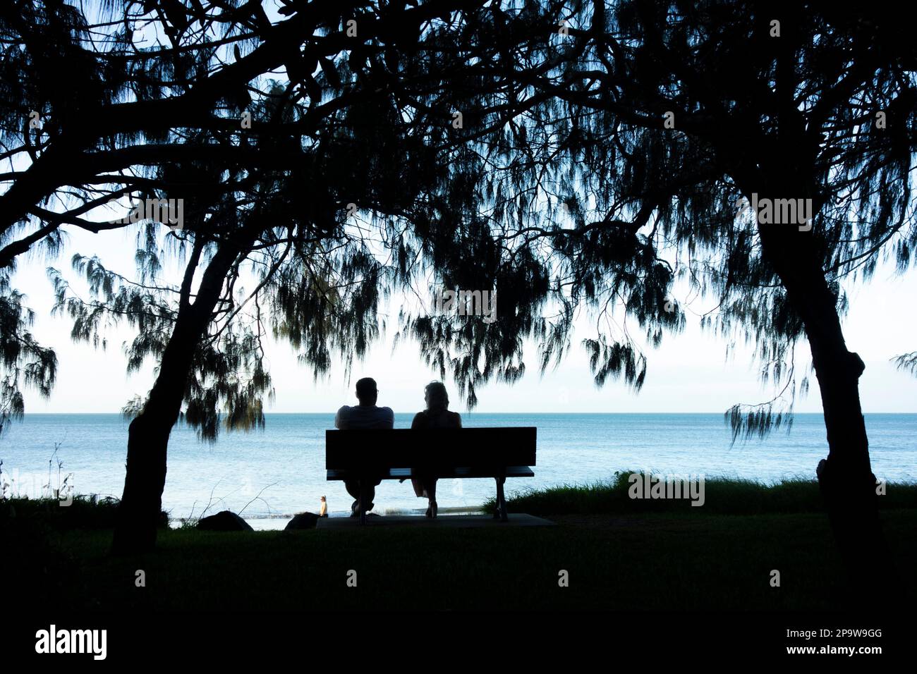 A rear view of a couple sitting on a wooden bench on the shore of Torquay beach, Hervey Bay, Queensland, Australia Stock Photo