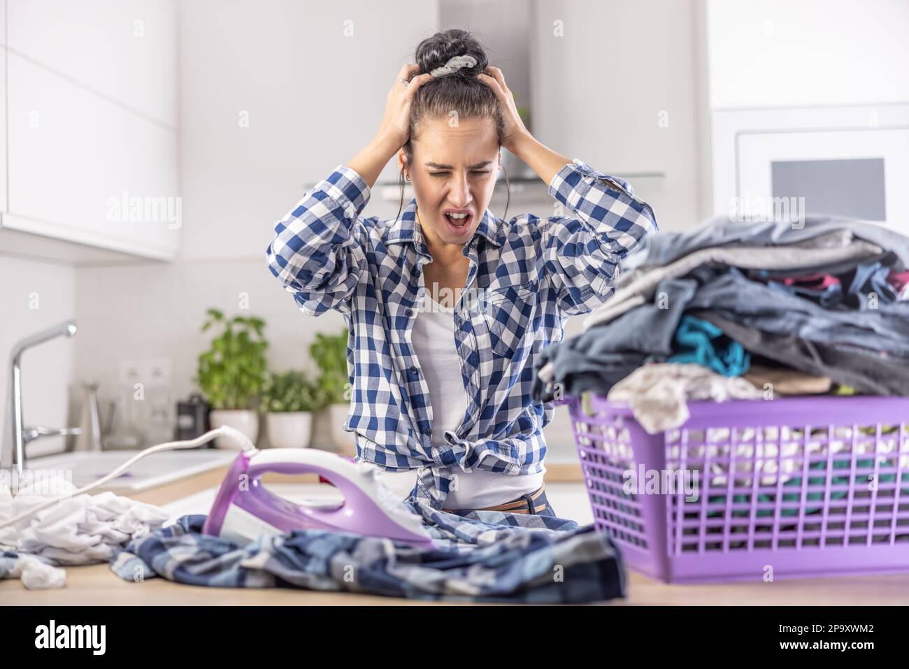 Woman shouts from anger and holds her head as she is frustrated from house chores and the pile of clothes she needs to iron. Stock Photo