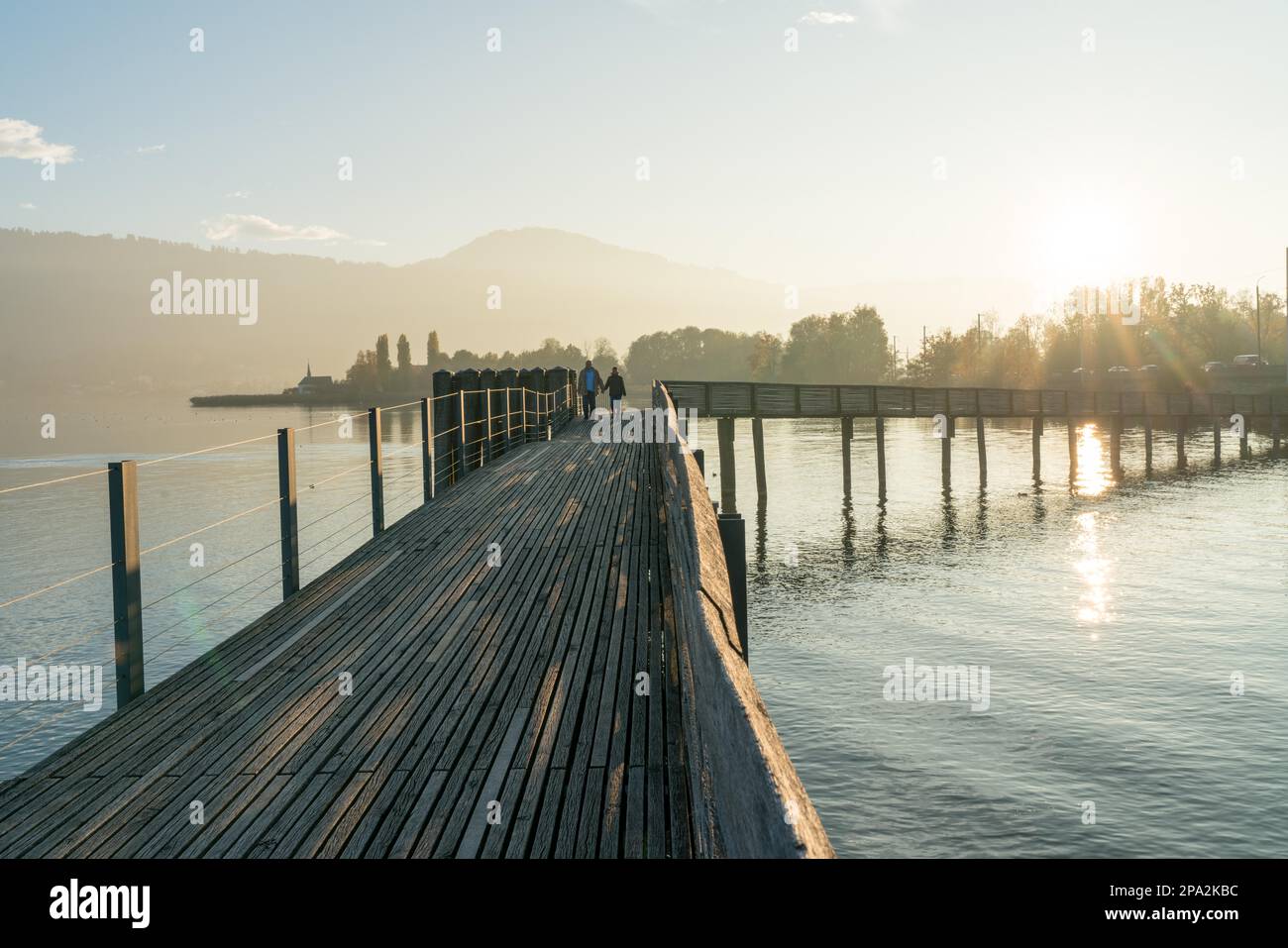 Landscape view of marsh and lake shore in evening light and a long wooden boardwalk in the foreground in late autumn in Switzerland with a couple Stock Photo
