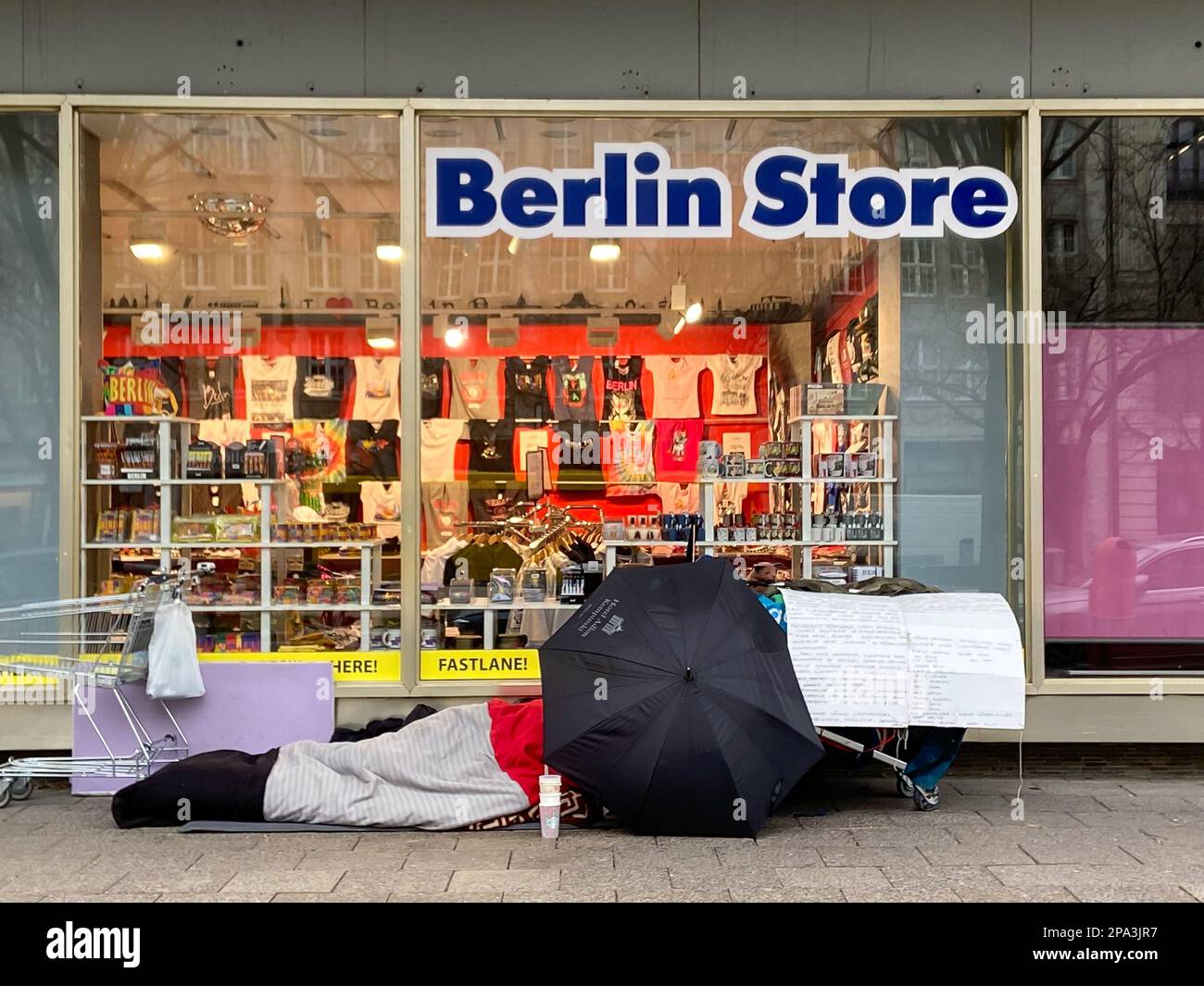 Homeless person in front of 'Berlin Store', covered by an umbrella from Hotel Adlon in central Berlin, Germany in February 2023. Stock Photo