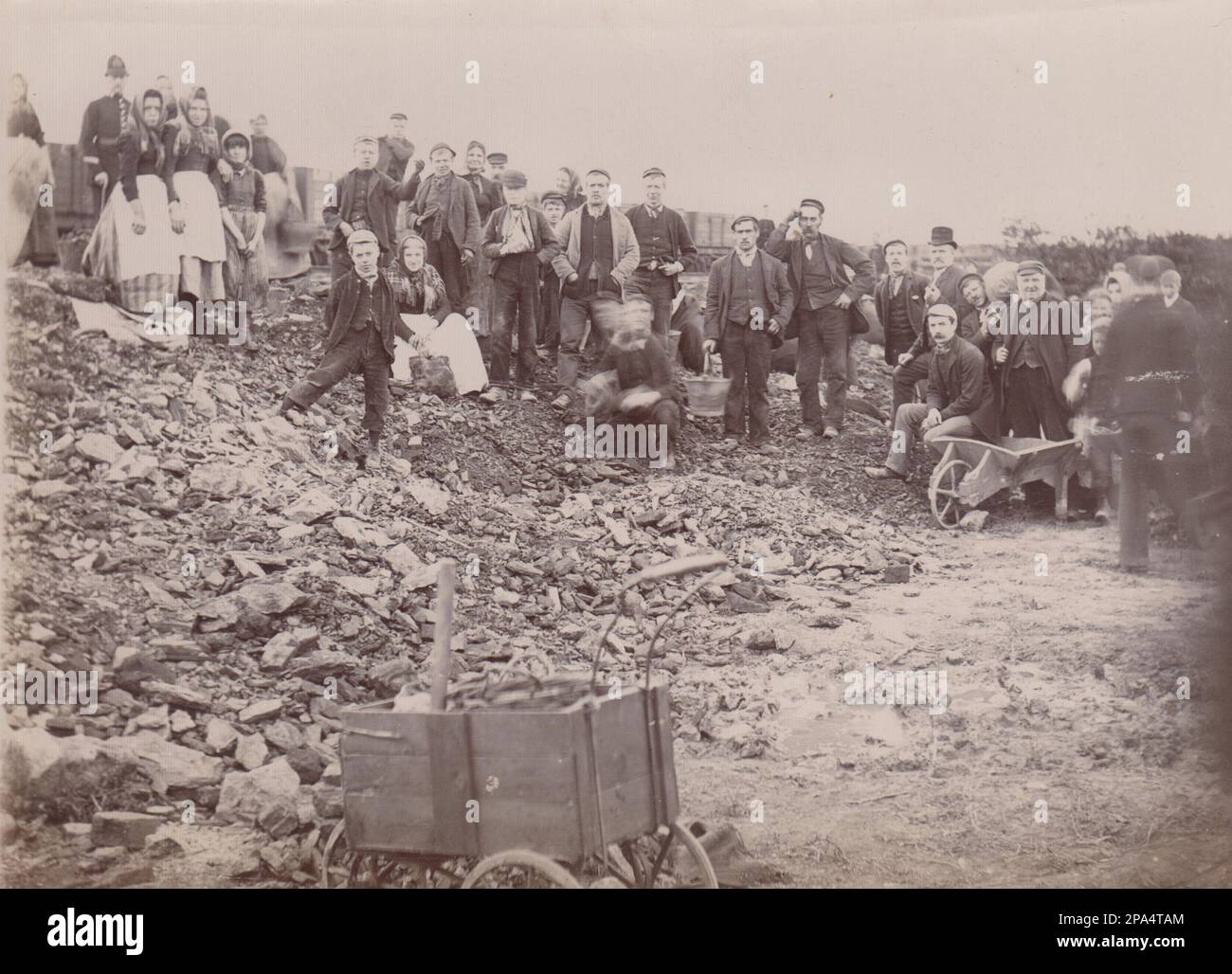 Families picking coal from a colliery soil heap. The early 20th century photograph shows men, women and children standing on the piled up waste material produced by the mine, they have paused from looking for coal to take home for fuel to look towards the camera. A policeman is at the back of the photo. The photograph is likely to have been taken during one of the early 20th century miners strikes Stock Photo