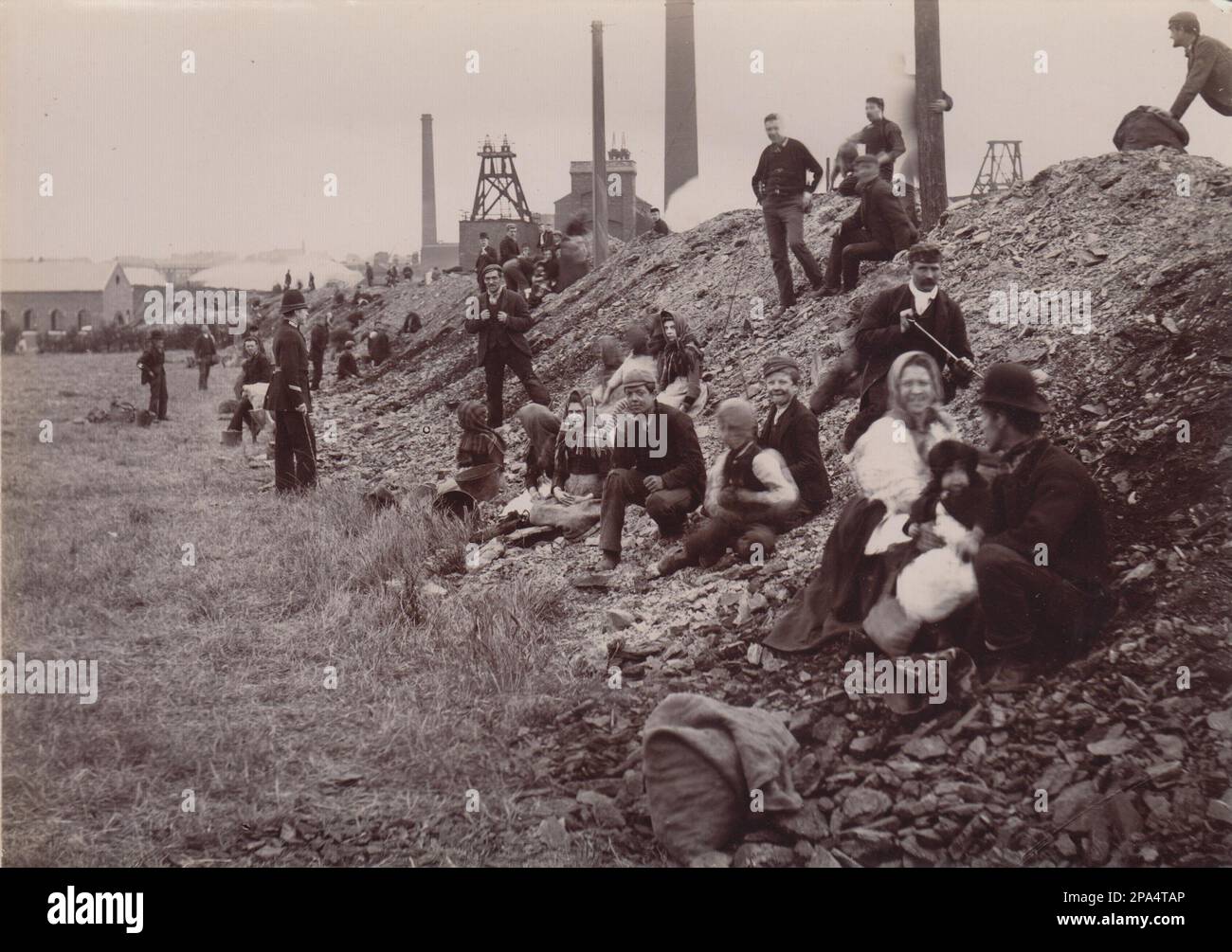 Families picking coal from a colliery soil heap. The early 20th century photograph shows men, women and children sorting through the waste material produced by the mine looking for coal to take home for fuel. Colliery buildings can be seen in the background, including a chimney and pit wheel. A policeman is watching the pickers. The photograph is likely to have been taken during one of the early 20th century miners strikes Stock Photo