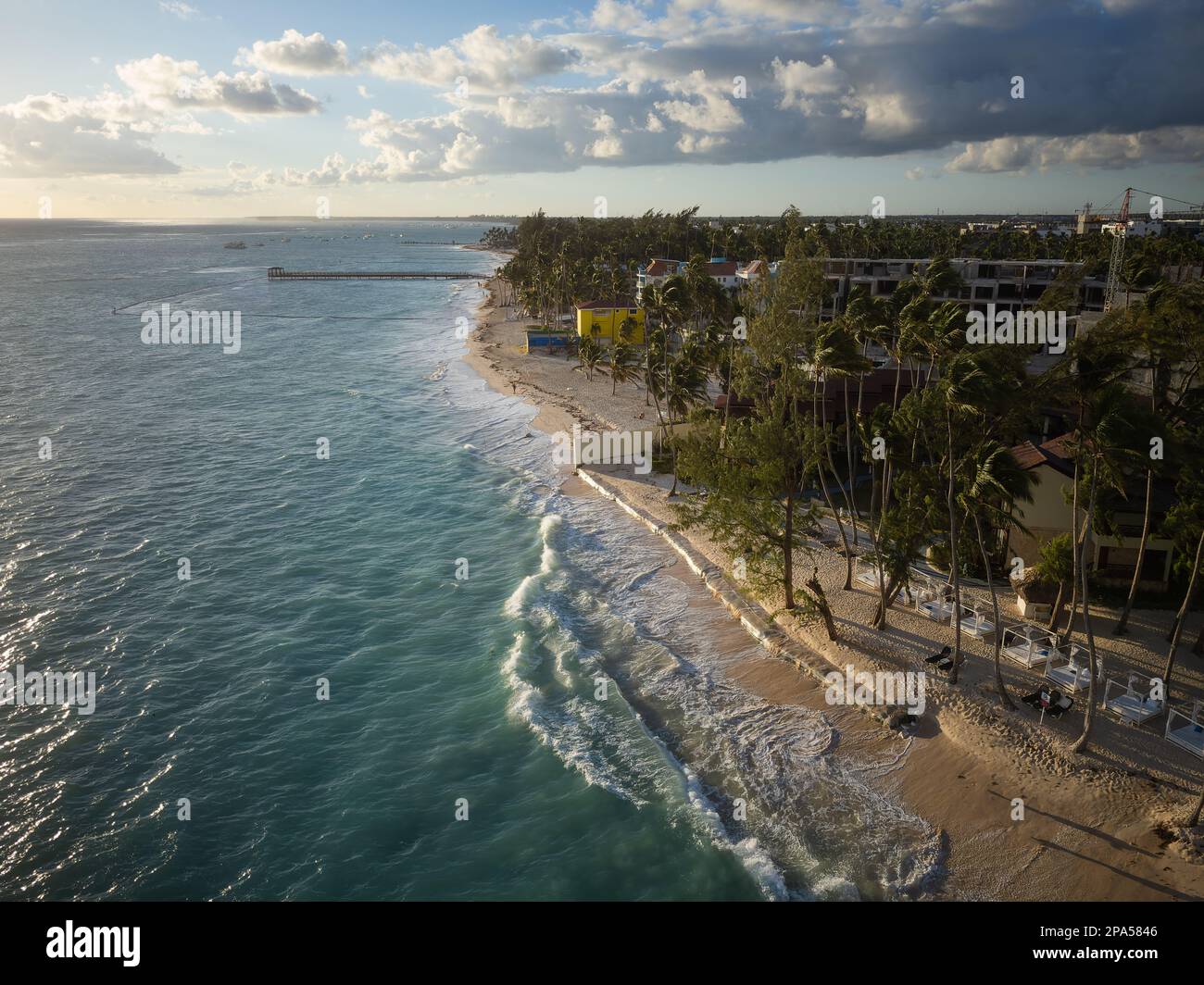 Shooting from a drone. A cozy resort town on the seashore. Clean sandy beach, tropical plants, turquoise sea water and white foamy waves. Cloudy sky. Stock Photo