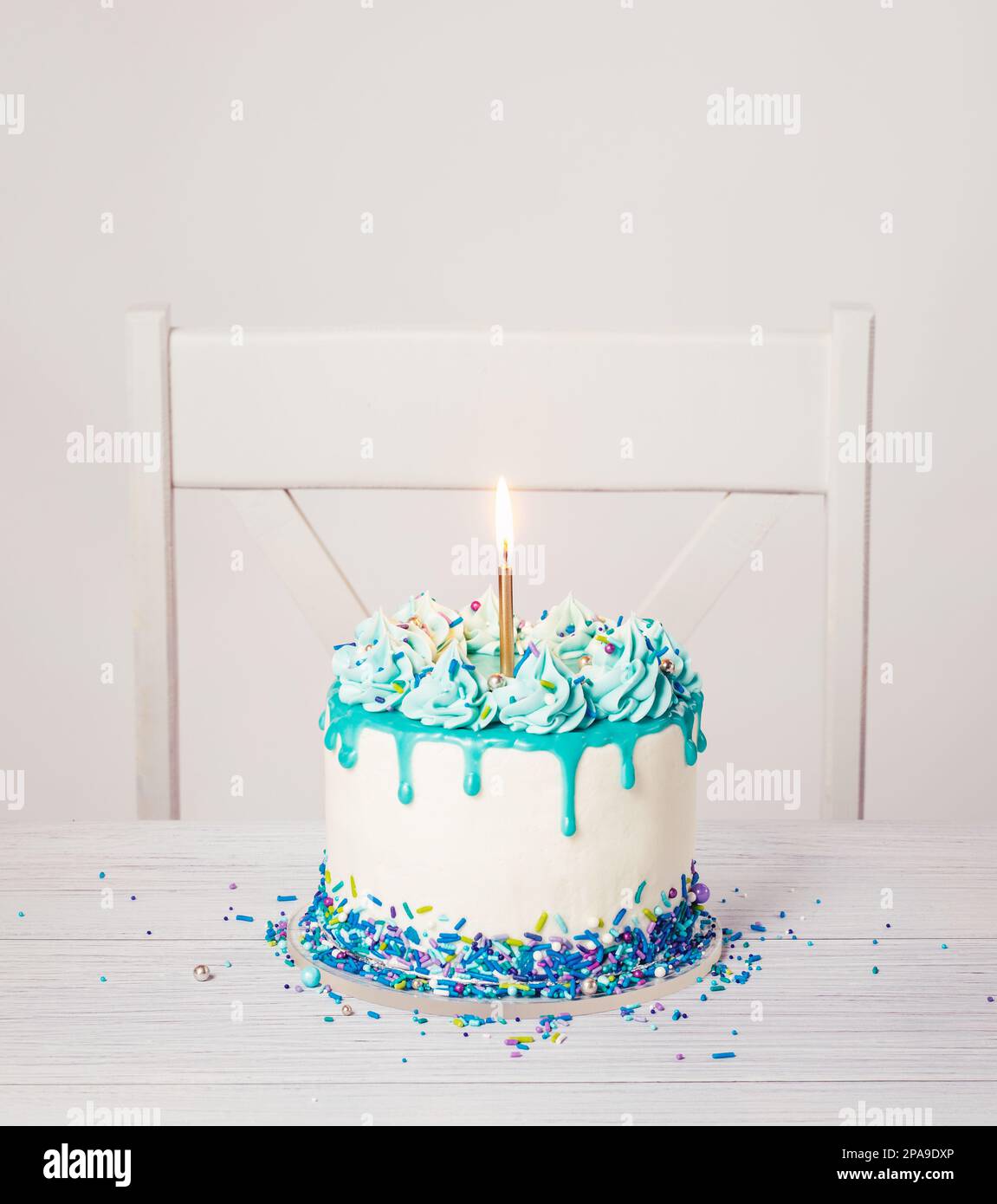 Blue and white buttercream birthday cake with a teal blue ganache drip, sprinkles, and bright gold candle on a white table and chair background. Trend Stock Photo