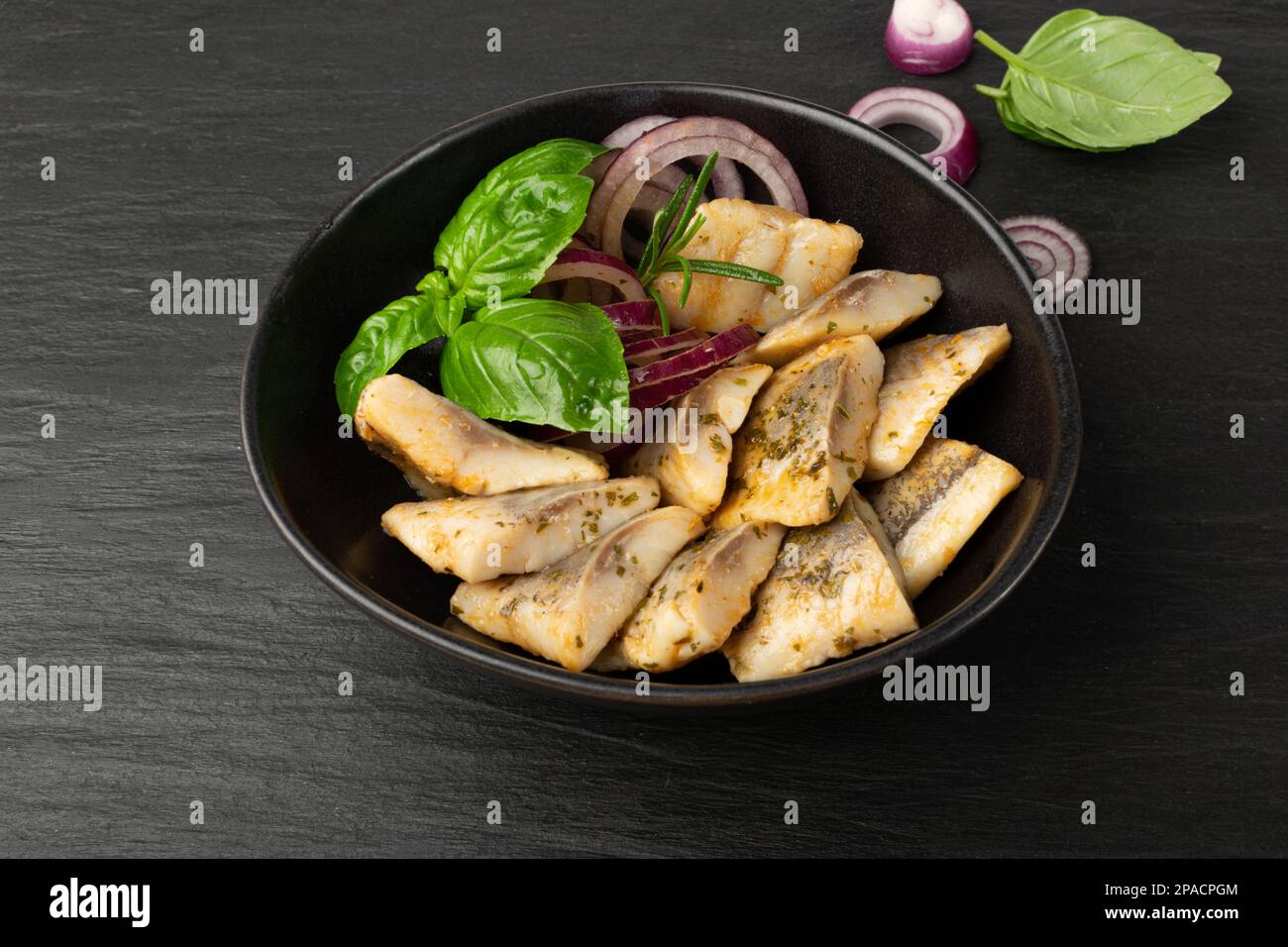 Salted Herring Fillet in Bowl, Raw Pickled Fish Meat with Herbs and Spices, Marinated Herring on Black Stone Background Stock Photo