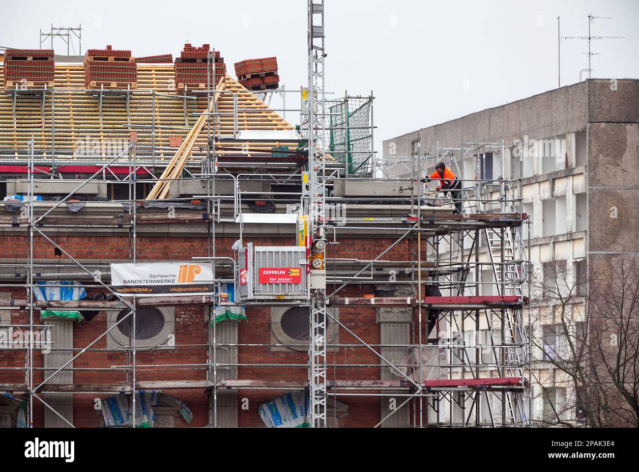 Potsdam, Germany. 10th Mar, 2023. A worker in orange protective clothing stands in the rain on the scaffolding of the future residential building Alter Markt No. 3. The roof tiles are already prepared on pallets for laying. In the background on the right, the GDR slab building Staudenhof can be seen. The new construction of an office and commercial building ensemble in the redevelopment area 'Potsdamer Mitte' was commissioned by the Potsdamer Wohnungsgenossenschaft 1956 eG. Credit: Soeren Stache/dpa/Alamy Live News Stock Photo