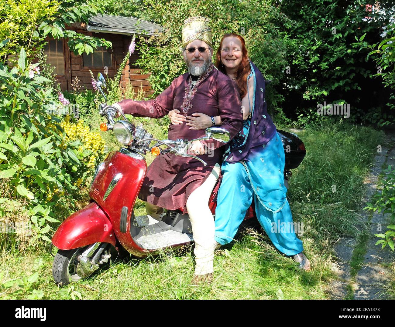 Just married European couple posing on a red electrical retro scooter in a garden. They are fan from Indian films Stock Photo