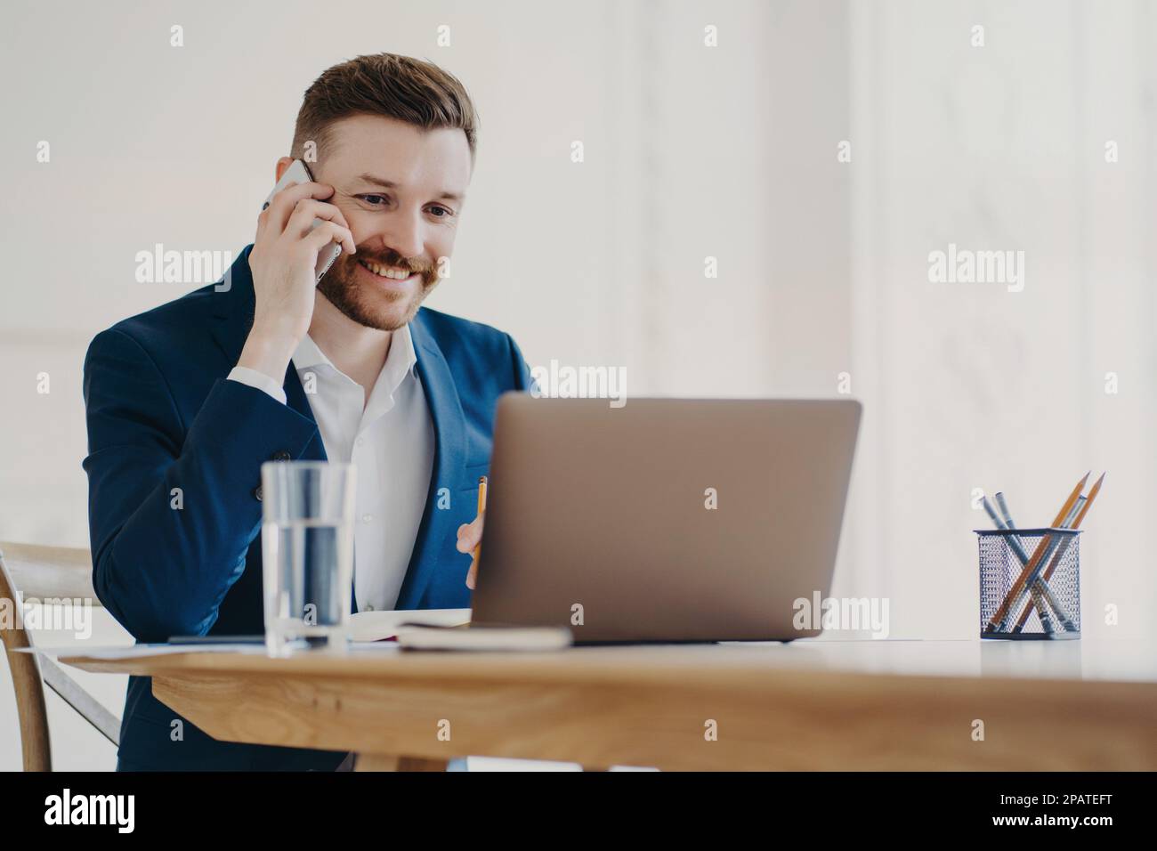 Young attractive businessman in formal suit smiling when talking on phone and looking at laptop screen approving what he sees, ceo sitting in minimali Stock Photo