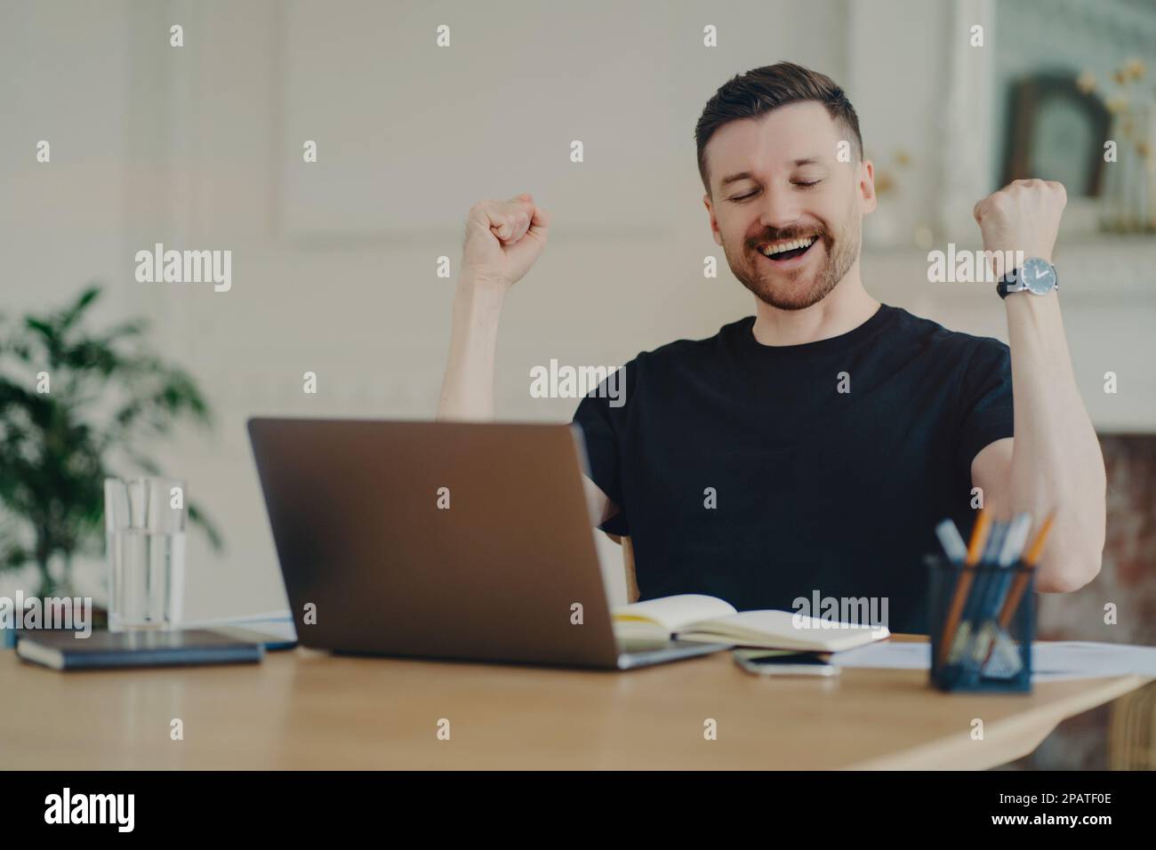 Excited cheerful bearded man with joyful expression clenches fists celebrates success dressed casually poses at desktop in front of opened laptop sati Stock Photo