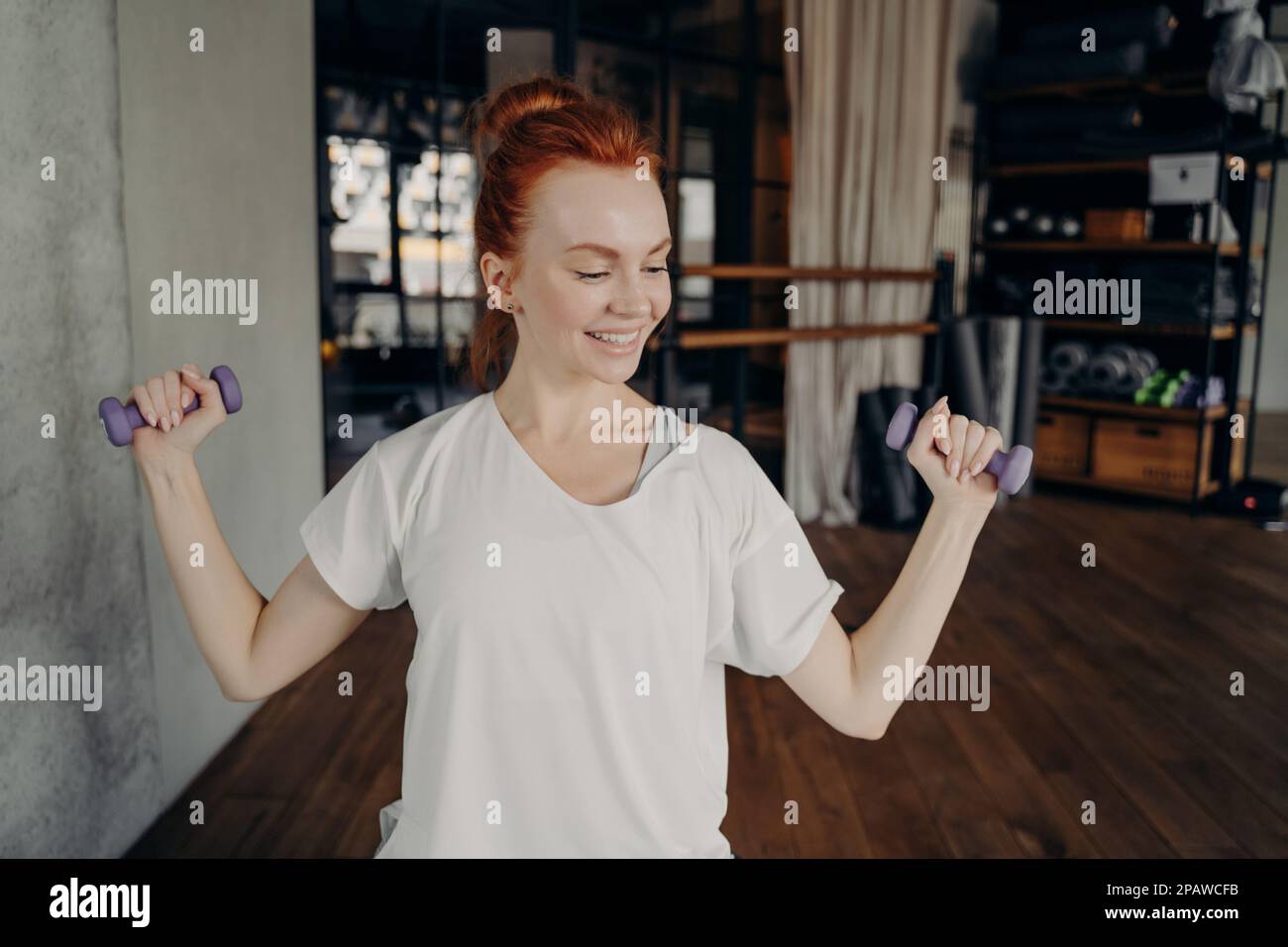 Healthy lifestyle. Young happy slim beautiful woman with red hair in bun working out in fitness studio, holding dumbbells and doing exercise for stron Stock Photo