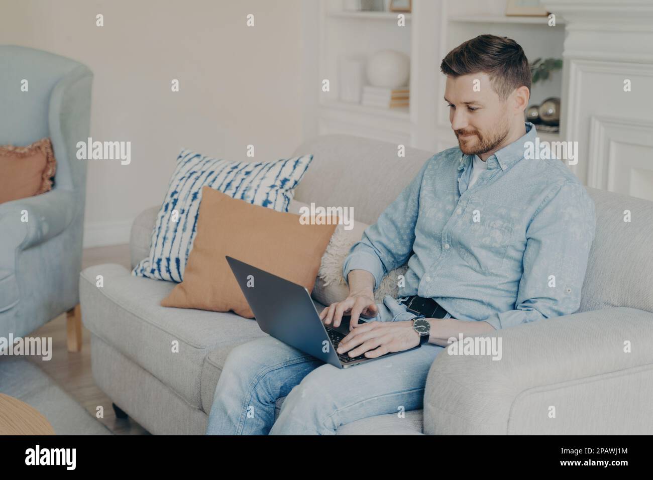 Relaxed casually dressed young businessman working from home while sitting on comfortable couch in light living room, typing on laptop and using speci Stock Photo