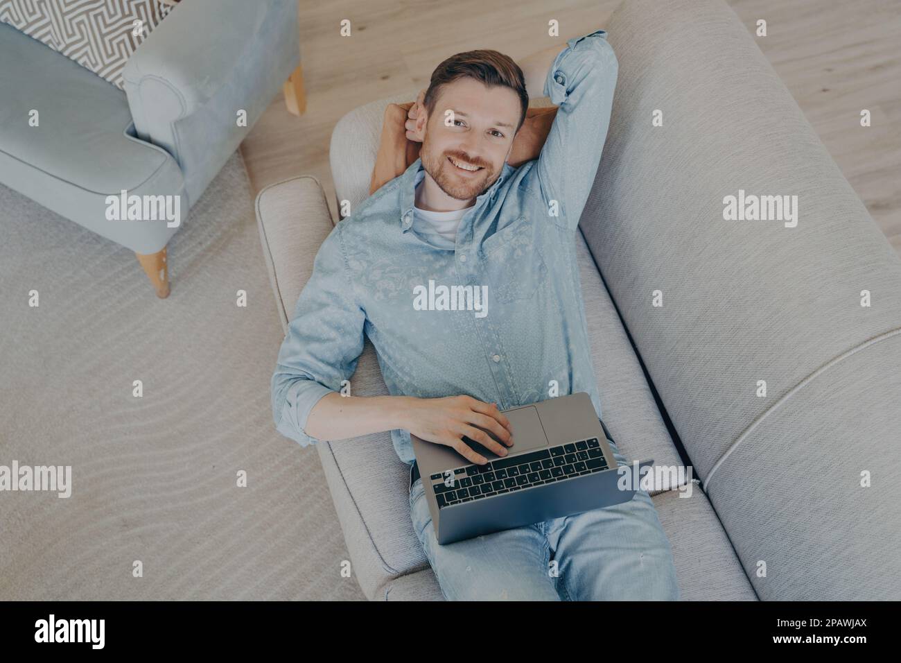 Happy young freelancer relaxing after finishing commission early, lying  on comfortable couch in living room, resting head on hand, looking up while s Stock Photo