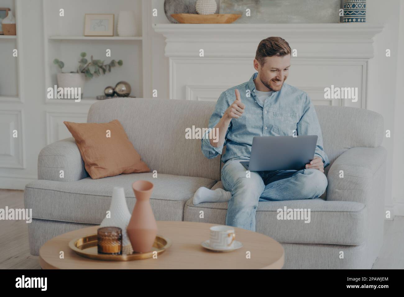 Young male business worker sitting on couch with crossed leg, showing thumbs up gesture to his co-workers in online meeting, displying  his satisfacti Stock Photo