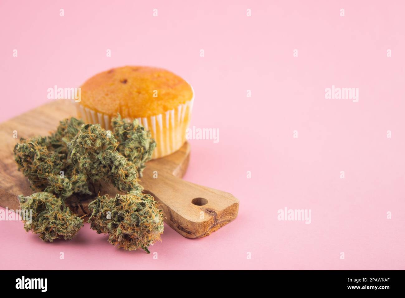 On a wooden board, a cupcake with the addition of cannabis, dry marijuana buds in the foreground.  On a pink background, a lot of empty space Stock Photo