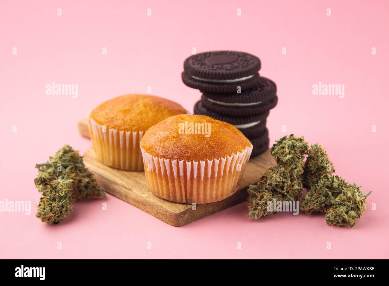 Cannabis cupcakes and chocolate chip cookies on a wooden board, dry marijuana buds on the sides.  On a pink background Stock Photo