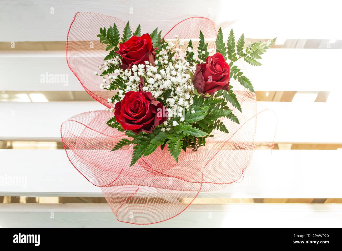 Three roses bouquet, with red wire mesh folds, on white horizontal wood boards, top view close up. Stock Photo